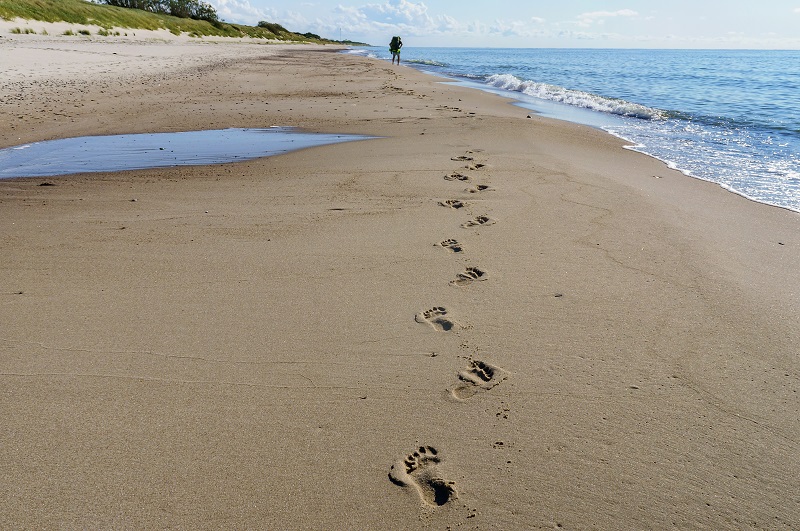 Nadmorska plaża z odciśniętymi śladami stóp, w oddali sylwetka człowieka