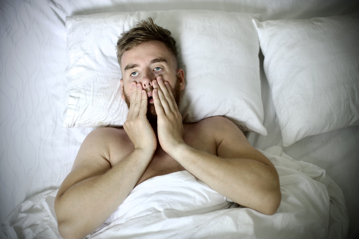 Młody mężczyzna w łóżku, obejmuje twarz dłońmi, smutny lub zmartwiony