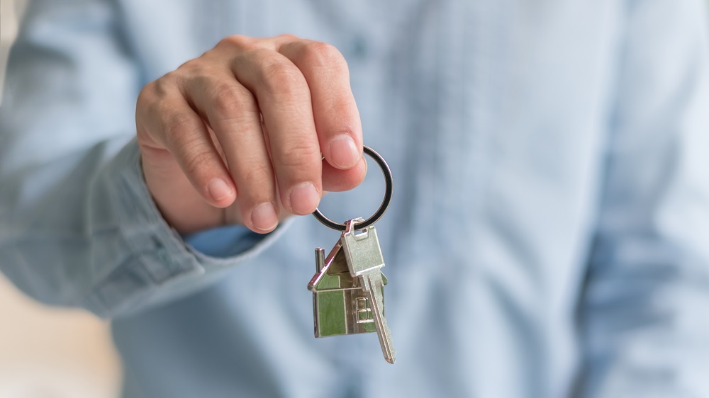 Mężczyzna trzyma przed sobą klucze z brelokiem w kształcie domu (kadr na sam tors i rękę z kluczami)