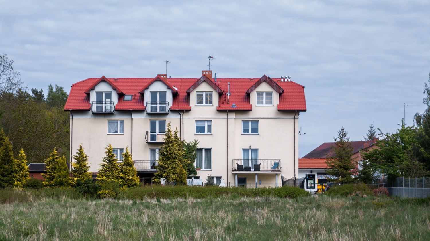 Niewielki blok mieszkalny z czerwonym, spadzistym dachem, otoczony zielenią i drzewami. Budynek ma balkony, jasną elewację oraz kilka kondygnacji, a w tle widać fragmenty innych zabudowań i niebo pokryte chmurami. Przed blokiem rośnie trawa i ozdobne krzewy.