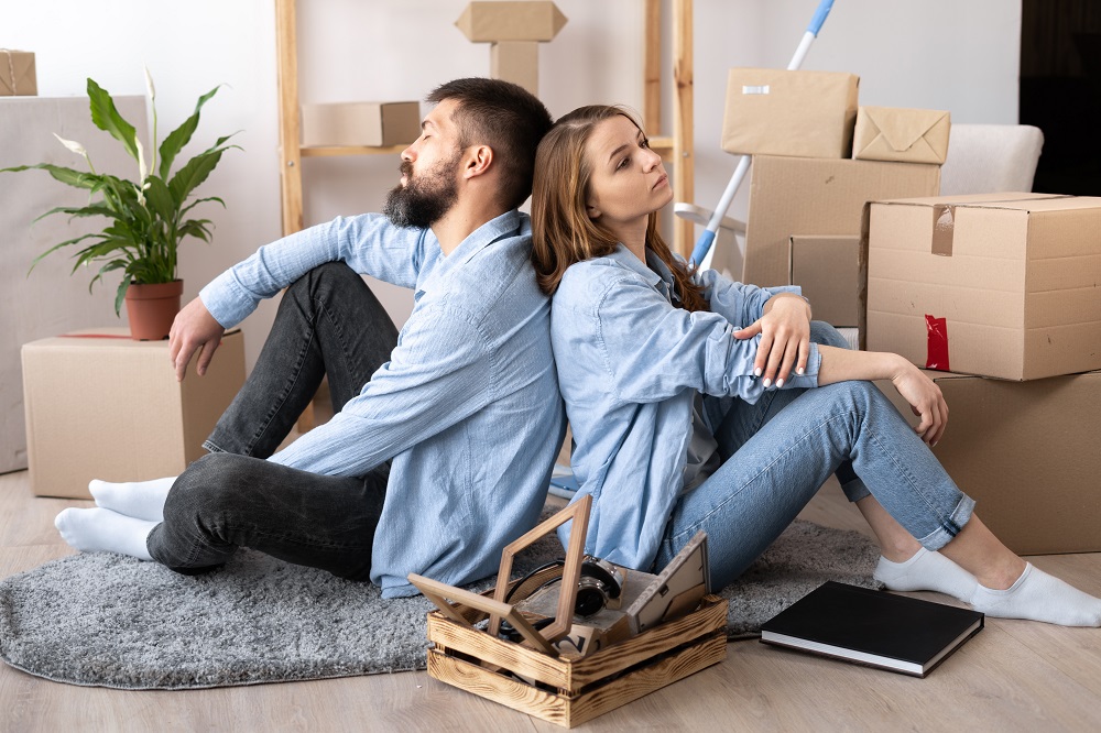 Młodzi kobieta i mężczyzna siedzą na podłodze, opierając się o siebie plecami, w nowym mieszkaniu, dookoła porozstawiane kartonowe pudełka