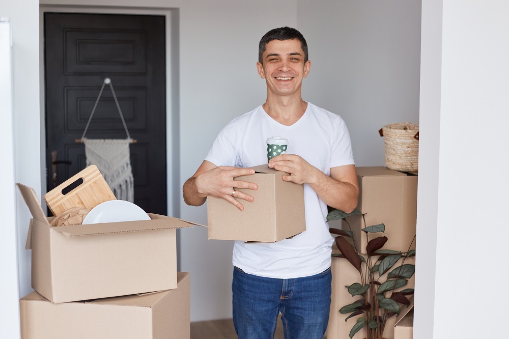Uśmiechnięty mężczyzna stoi z kartonowym pudełkiem w pomieszczeniu, dookoła sterty pudełek ze spakowanymi rzeczami