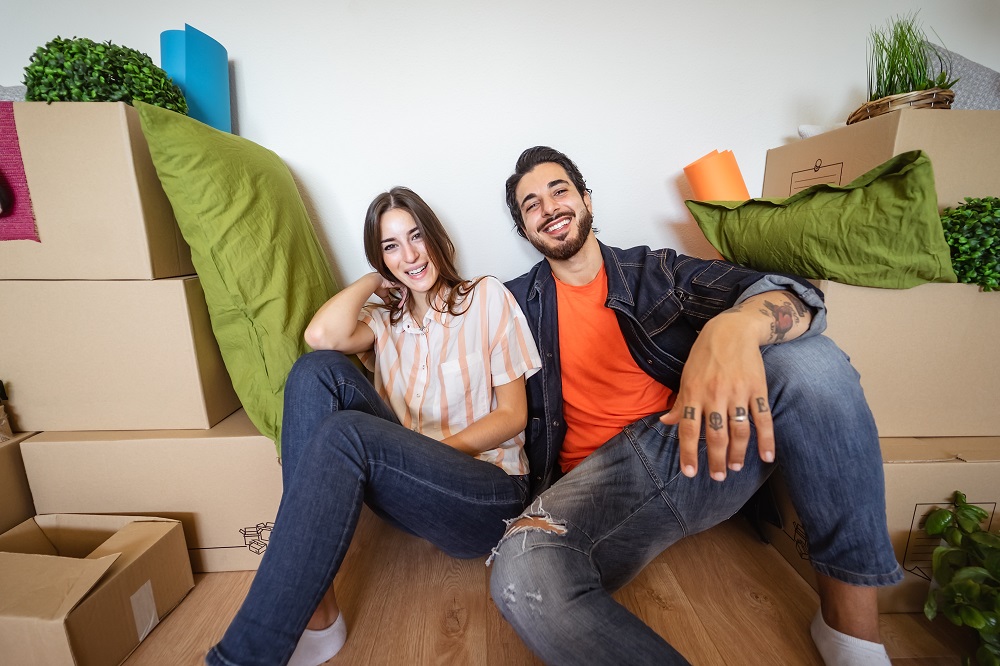 Uśmiechnięta młoda para siedzi pod ścianą nowego mieszkania, otoczeni nierozpakowanymi rzeczami w pudełkach