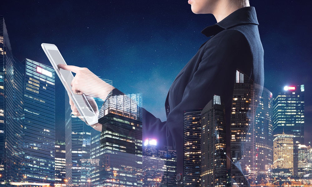 Kobieta w biznesowym stroju pracuje na tablecie, na nią naniesiona półprzezroczysta panorama nowoczesnej metropolii nocą