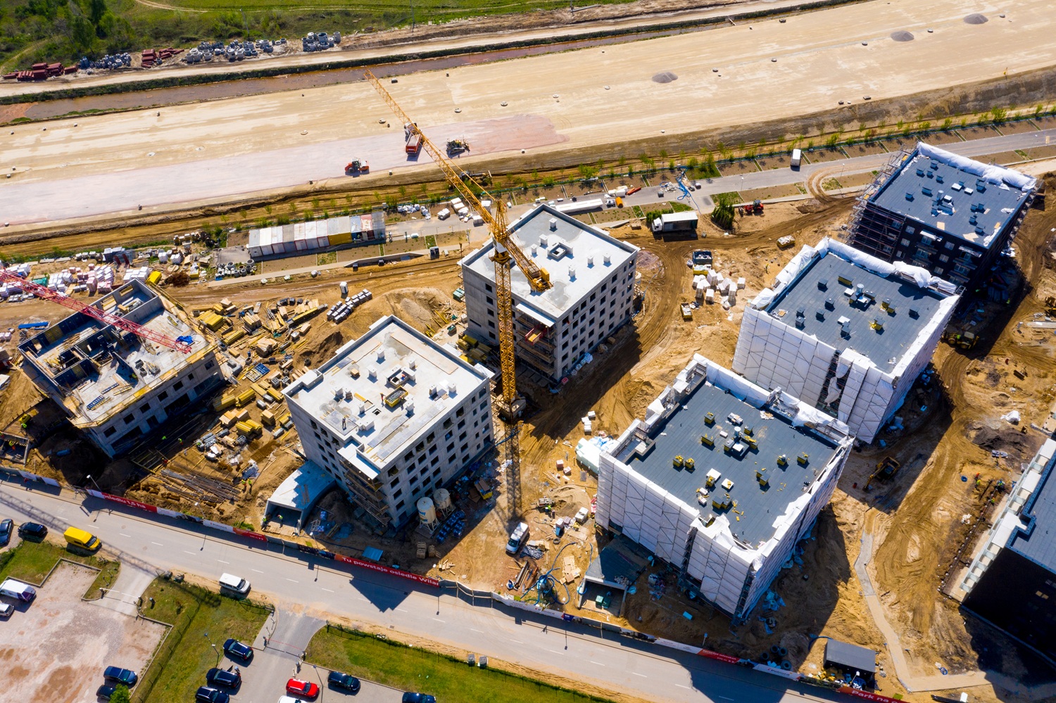 Zdjęcie lotnicze przedstawia plac budowy osiedla mieszkaniowego. Widać kilka nowo powstających bloków, żurawie budowlane, materiały budowlane oraz pracujący sprzęt. Część budynków jest w stanie surowym, inne są już częściowo wykończone lub osłonięte folią. Całość otoczona jest drogą i zaparkowanymi samochodami.