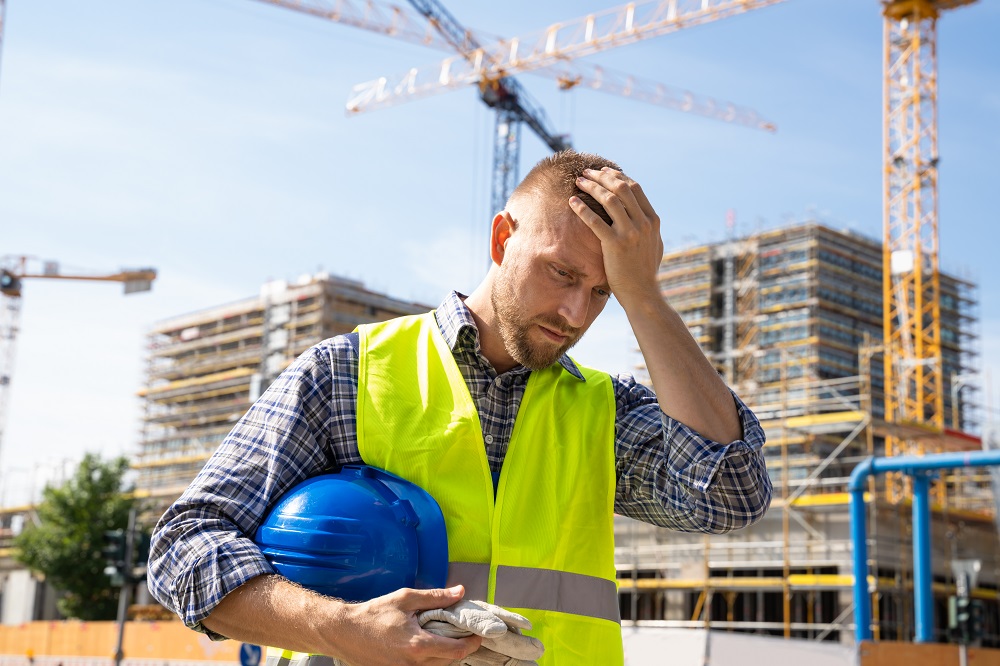 Mężczyzna w stroju robotnika (odblaskowa kamizelka, kask) trzyma się za głowę, w tle teren budowy bloków mieszkalnych