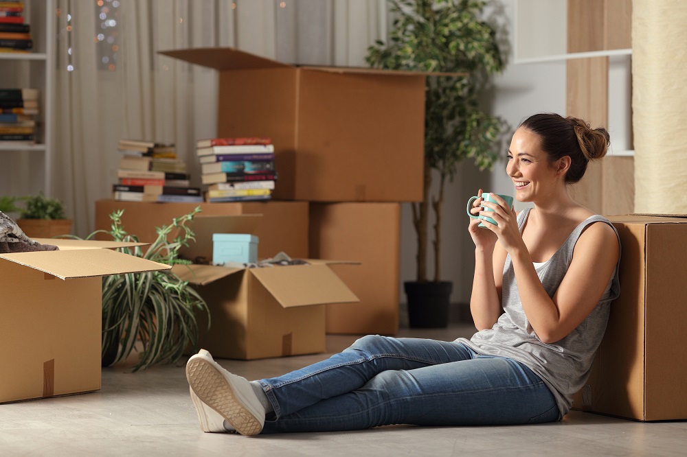 Młoda uśmiechnięta kobieta siedzi na podłodze z kubkiem, w tle kartonowe pudełka z rzeczami z przeprowadzki