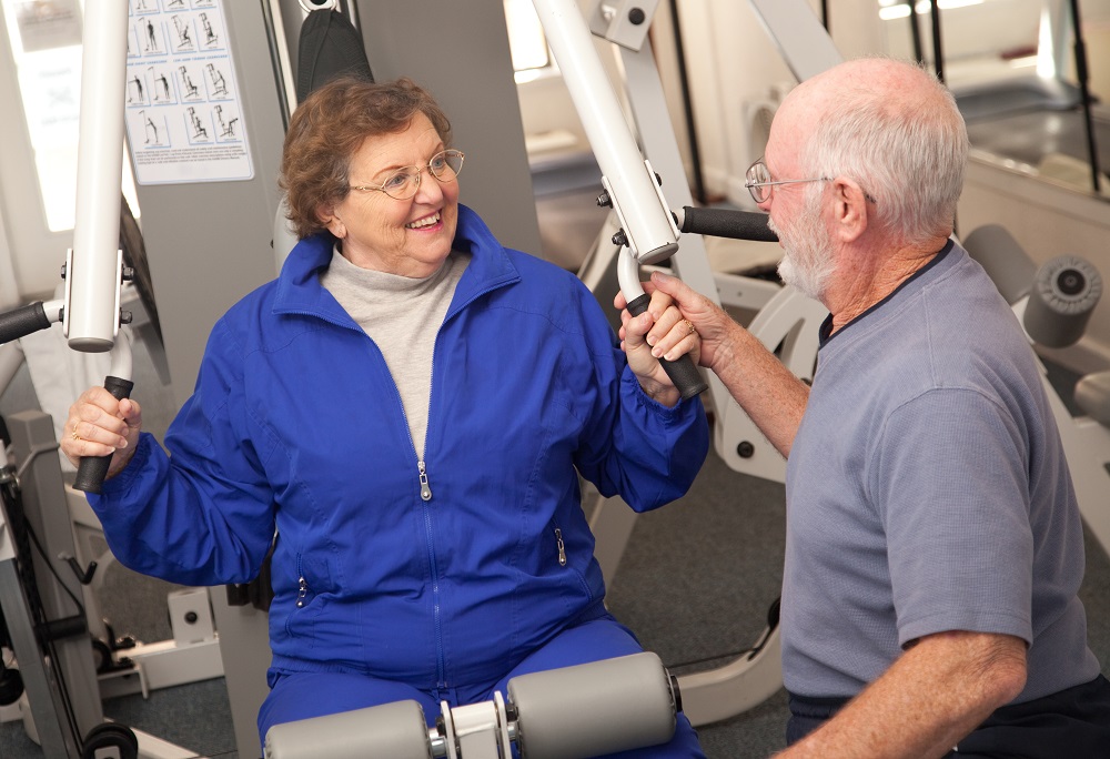 Uśmiechnięci starzy ludzie - kobieta ćwiczy na urządzeniu na siłowni, a mężczyzna siedzi obok